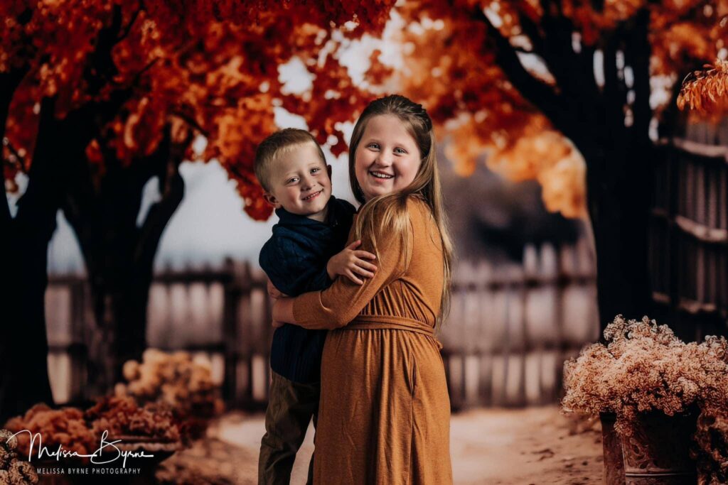 Brother and sister hug each other in front of trees with Fall colors in Fall mini photo session.